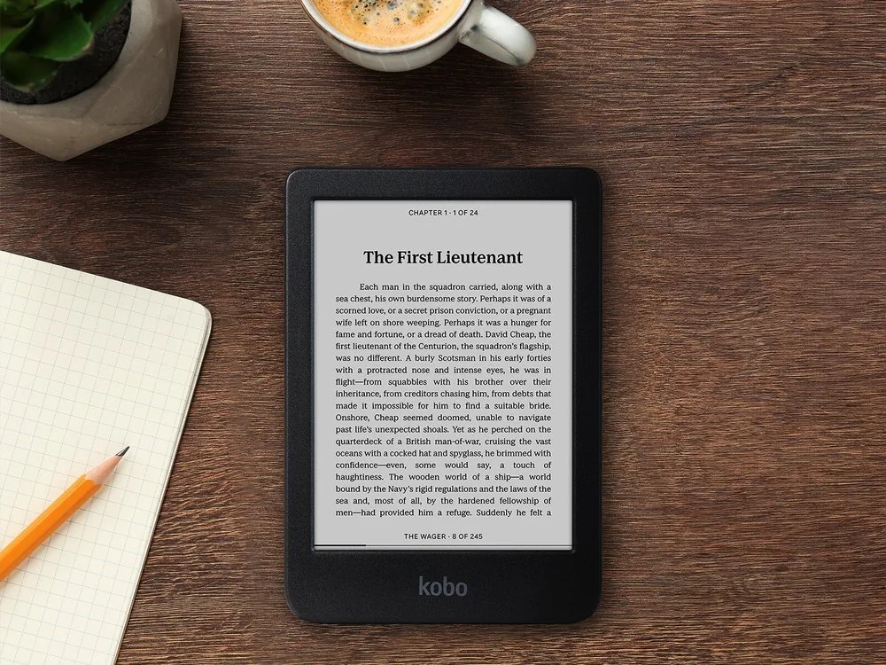 Un Kobo Clara BW con la página de un libro, sobre una mesa de madera junto a un café y una libreta